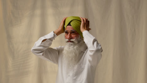 Fotografía-De-Estudio-De-Un-Hombre-Sikh-Mayor-Que-Se-Arregla-La-Barba-Y-El-Bigote-Mientras-Se-Pone-Un-Turbante-Contra-Un-Fondo-Liso